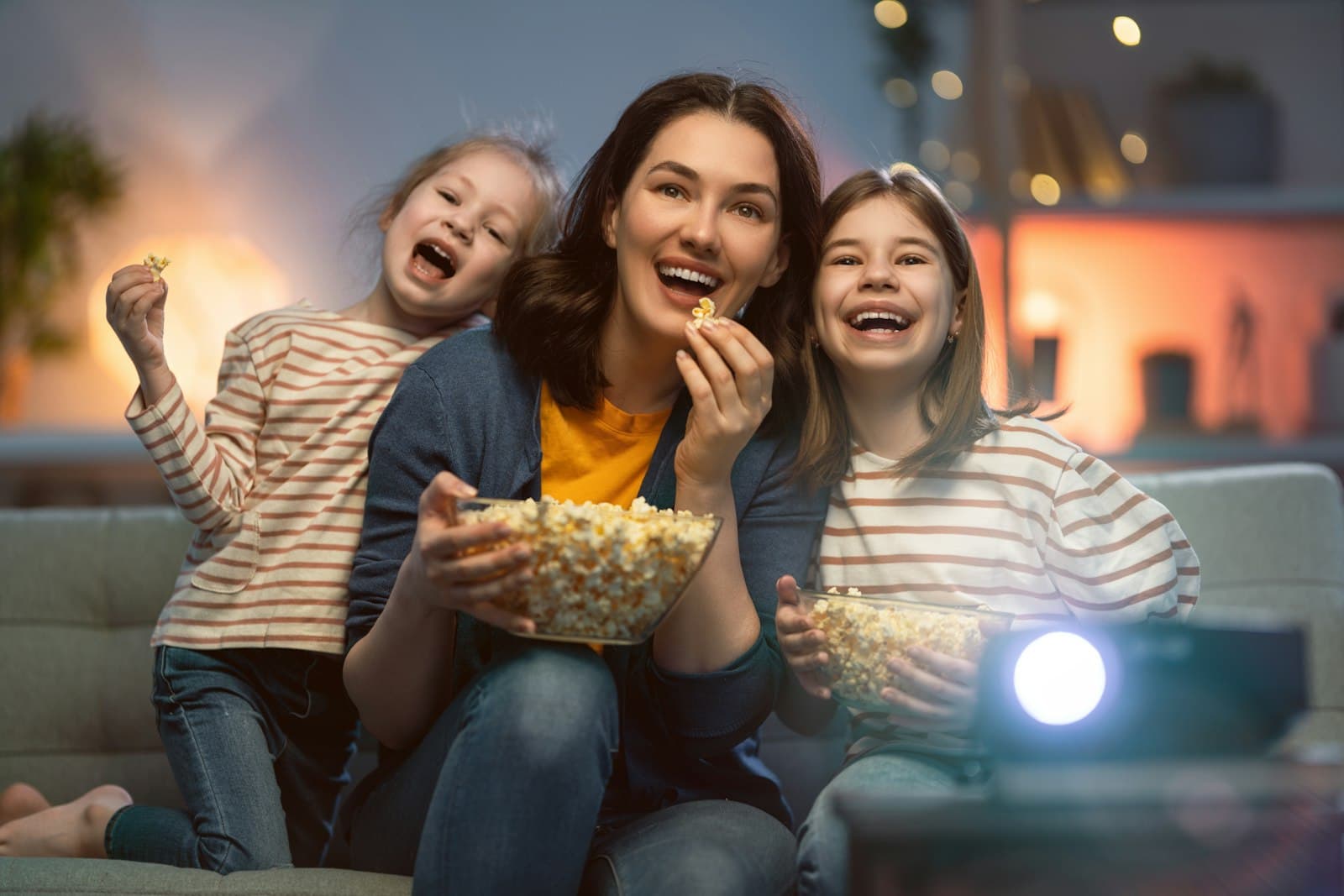 Une mère et ses deux filles rient sur le canapé en regardant un film, un bol de pop-corn à la main